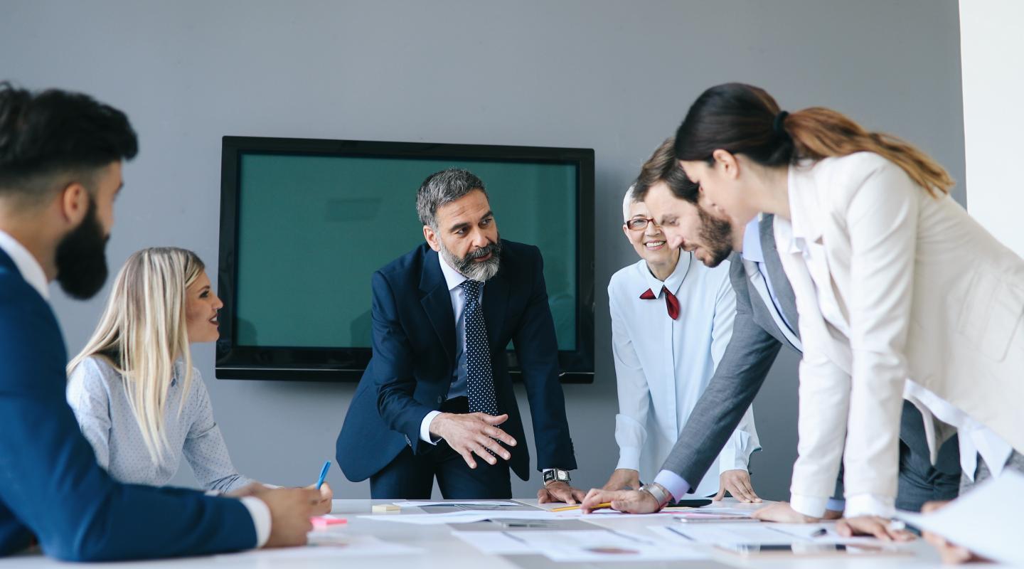 Three male and three female office workers standing and gathered around a table speaking to each other