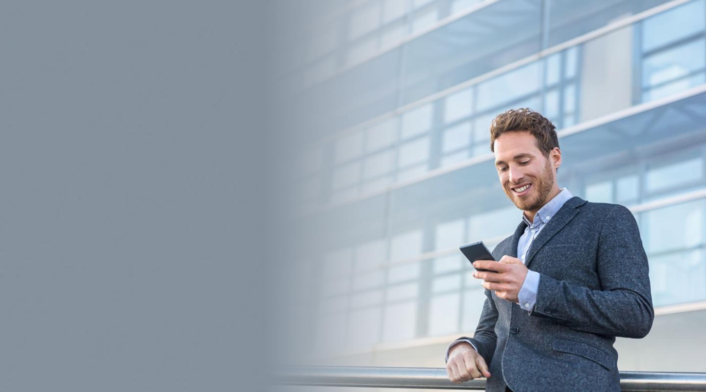 Young male in business suit smiling and looking at his phone, standing in front of an office building on the right side of the image.