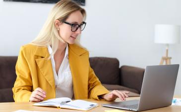 young woman with blonde hair and glasses wearing a yellow business jacket and white shirt working on her laptop