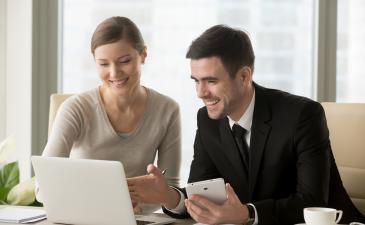 A young man and woman in an office both on a laptop smiling, collaborating
