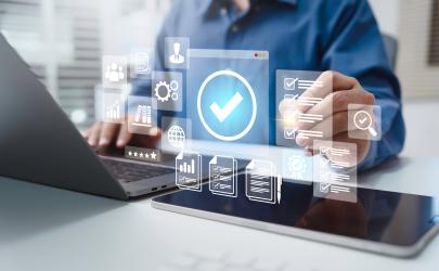 Male professional working on laptop and tablet pointing to a certified checkmark graphic. Other icons showing technology, documents, people, and graphs surround the check mark.
