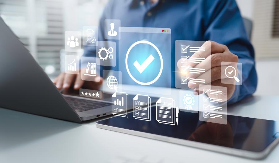 Male professional working on laptop and tablet pointing to a certified checkmark graphic. Other icons showing technology, documents, people, and graphs surround the check mark.